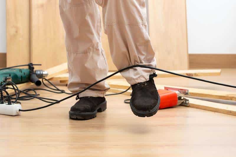 A worker is about to trip on an electrical cord - a common cause for trip and fall incidents at work. Learn more at 1stReporting.com.