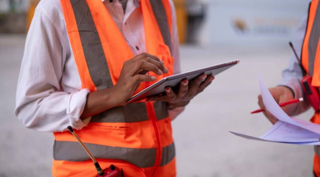 A safety officer adds a safety document to digital format using a tablet while a second safety officer dictates from a paper version what the digital tailboard meeting form must include. Learn more about digital tailboard meeting form use at 1stReporting.com.