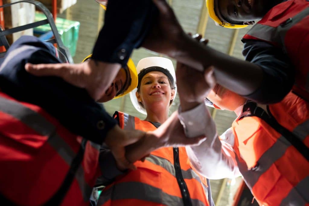 A group of workers do a motivational meeting handshake during a tailboard meeting.