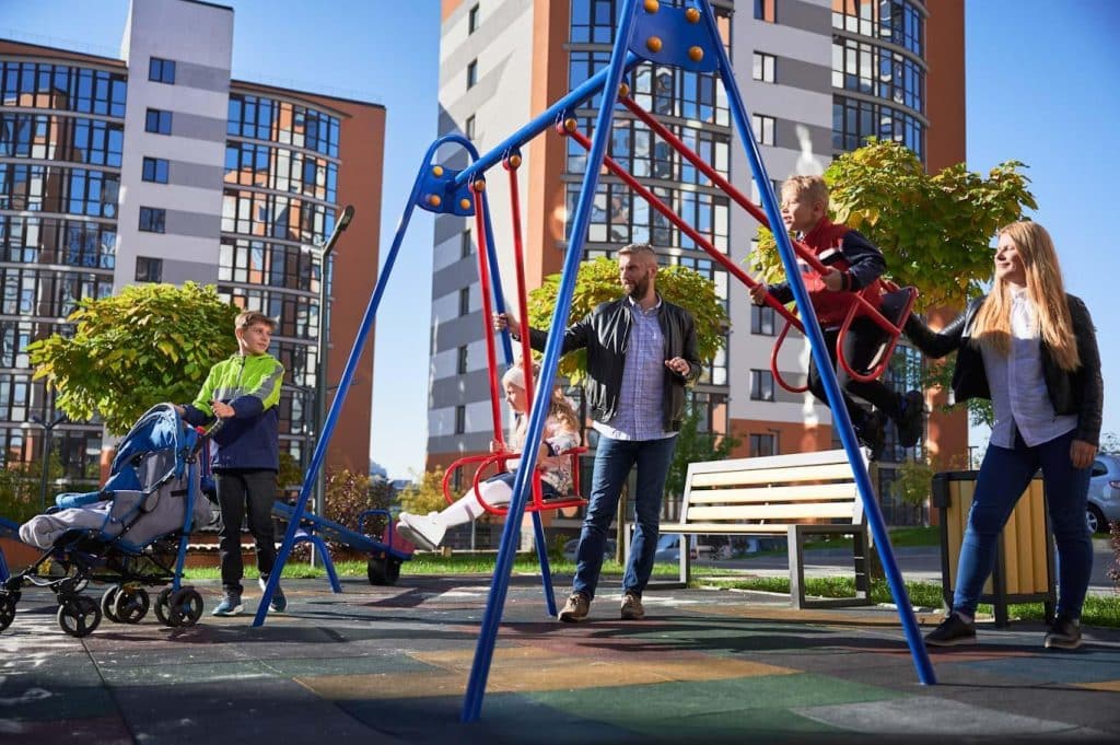 A family playing on a public swing set in a playground. Learn about playground safety audit form use at 1stReporting.com.